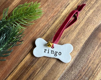 May include: A white, bone-shaped pet tag with the name "ringo" in black lettering. It has a small gold bell and a burgundy ribbon for hanging. The tag is set against a wooden background with a green sprig.
