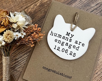 May include: A white cat-shaped tag with the text "My humans are engaged 12.06.25" hangs from a twine string. It rests on a brown card with "congratulations!" printed below. A small bouquet of dried flowers is to the left.