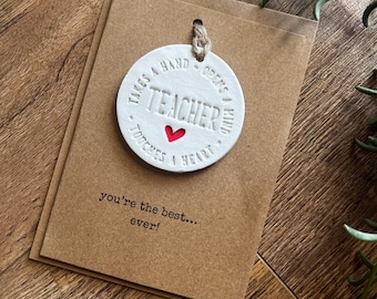 May include: A white ceramic teacher gift tag with the words "TEACHER" and "Takes a hand, opens a mind, touches a heart" and a red heart. The tag is attached to a brown card that reads "you're the best... ever!"