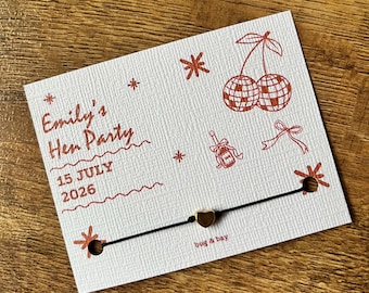 May include: A white card with red text and illustrations, including cherries and a champagne bottle. The card reads "Emily's Hen Party 15 July 2026." A black string with a gold heart charm is attached.