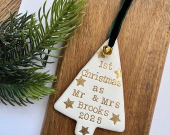 May include: White ceramic Christmas tree ornament with gold text and star accents. The ornament reads "1st Christmas as Mr & Mrs Brooks 2025." It has a green velvet ribbon and a small gold bell for hanging.