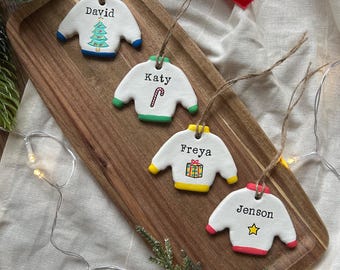 May include: Four white ceramic sweater ornaments with colorful trim and names: David, Katy, Freya, and Jenson. Each ornament has a unique design, including a Christmas tree, candy cane, gift, and star. They hang from twine strings.