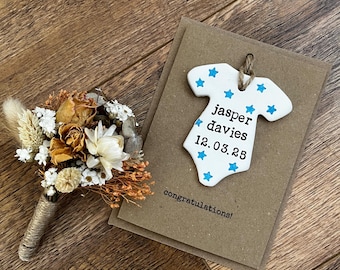 May include: A dried flower boutonniere with a twine-wrapped stem rests on a wooden surface. A white onesie-shaped ornament with blue star accents and the name "Jasper Davies" and date "12.03.25" is next to a card that says "congratulations!"