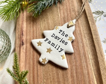 Könnte beinhalten: Weißer Weihnachtsbaumschmuck aus Keramik mit goldenen Sternen und dem Text "The Davies Family". Der Anhänger hängt an einer Schnur aus Naturfaser. Auf einem Holztablett mit grünen Zweigen.