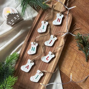May include: Seven white ceramic Christmas stocking ornaments are arranged on a wooden serving board. Each stocking features a letter in red or green, with red and green dot details. The ornaments are suspended by twine.