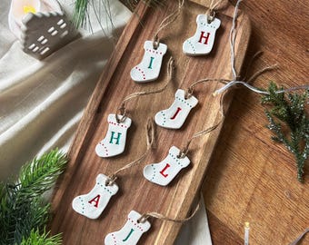 May include: A wooden serving board displays seven white ceramic Christmas stocking ornaments. Each stocking has a letter in red or green, with red and green dot accents. The ornaments hang from twine.