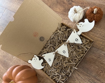 May include: A Halloween-themed gift box with a "BOO" banner, ghost-shaped ornaments, and decorative pumpkins. The banner is made of white triangles with black letters. The box is filled with shredded paper.