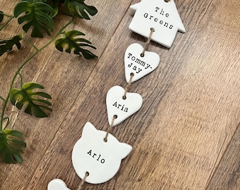 May include: A white ceramic family and pet name tag set. The set includes a house, heart, cat, and bone shapes, each with a name printed on it: "The Greens", "Tommy-Jay", "Aria", "Arlo", and "Buddy". The tags are strung together with twine.