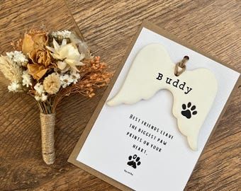 May include: A dried flower boutonniere with a twine-wrapped stem sits next to a card with a wooden wing ornament. The card reads "Buddy" and "Best friends leave the biggest paw prints on your heart."