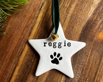 May include: White star-shaped ornament with the name "reggie" in black text and a black paw print. It has a green ribbon and a small gold bell for hanging. The ornament is on a wooden surface.