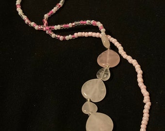 Puede incluir: Un delicado collar con una hebra de pequeñas cuentas rosas y blancas. El collar tiene una serie de piedras más grandes, en forma de corazón, de color rosa pálido. El collar se muestra sobre un fondo negro.