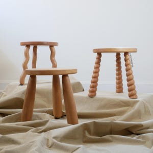 Puede incluir: Dos taburetes de madera clara con asientos redondos. Un taburete tiene tres patas lisas y cónicas, mientras que el otro presenta tres patas retorcidas en forma de cuerda. Ambos taburetes están hechos de madera natural con veta visible.