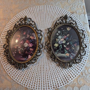 May include: Two oval wall frames with ornate, antique-style bronze-colored metal borders. Each frame contains a dark, detailed print of a floral bouquet featuring various colorful flowers, including pink, white, and yellow blossoms against a dark background.