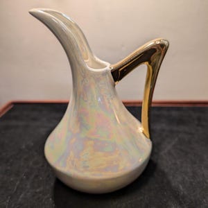 May include: Iridescent ceramic pitcher with a gold handle. The pitcher has a pearlescent finish that reflects a rainbow of colors. The handle is a striking gold color, adding a touch of elegance to the piece. The pitcher is likely used for serving beverages.