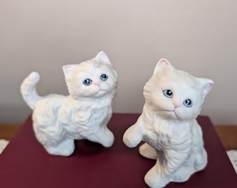 Puede incluir: Dos figuras de gato de cerámica blanca con pelaje texturizado esponjoso, ojos azul brillante y narices rosadas. Un gato está de pie con la cola levantada, mientras que el otro está sentado con una pata ligeramente levantada. Detalles faciales.