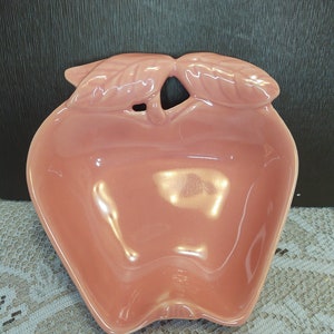 May include: A pink ceramic apple-shaped dish with a leaf design on the top. The dish is perfect for serving snacks or holding small items.