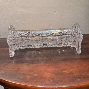 May include: A clear, rectangular glass dish with a cut-glass pattern. The dish has a decorative, openwork design on the sides and ends, and is elevated on four small feet. The dish is likely for serving or display.