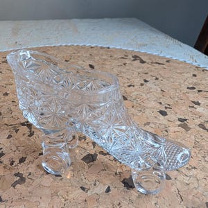 May include: A clear glass figurine shaped like a roller skate with a detailed starburst pattern pressed into the surface. The skate features a high-top boot design with a defined heel and toe area, supported by two circular glass wheels at the base.