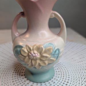 May include: A ceramic vase featuring a soft pastel color palette of pink, cream, and light blue. The vase has two curved handles, a flared rim, and a raised, three-dimensional floral relief design on the front center with light blue leaves.