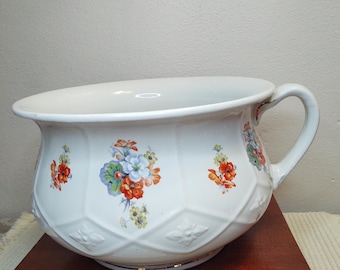 May include: A white ceramic chamber pot with a floral design. The pot has a handle and a gold rim. It is sitting on a wooden surface.