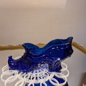 May include: A cobalt blue glass shoe-shaped decorative piece with ornate detailing. The shoe sits on a white, circular, interlocking ring design. The glass has a glossy finish, reflecting light. The shoe's design is reminiscent of vintage decor.