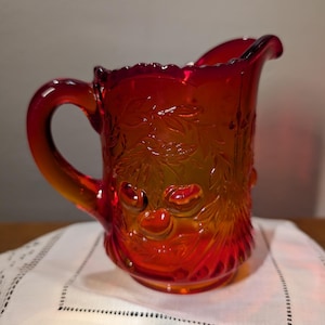 May include: A vibrant red glass pitcher with a decorative floral pattern. The pitcher has a curved handle and a ruffled rim. The glass has an orange hue in the center. The pitcher is sitting on a white, embroidered cloth.