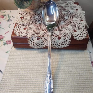 May include: A silver serving spoon with an ornate handle. The spoon is on a white crocheted doily.