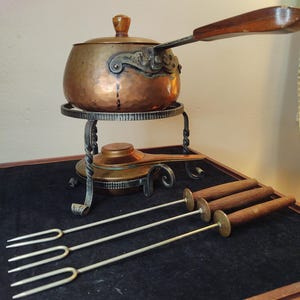May include: A vintage copper fondue pot with a wooden handle and four metal forks. The pot is sitting on a metal stand with a burner. The set is on a dark brown wooden surface.