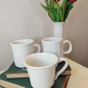 May include: Three white ceramic mugs with handles, two are plain and one has a textured design. The mugs are stacked on top of two books with a green cover. A white vase with red tulips is in the background.