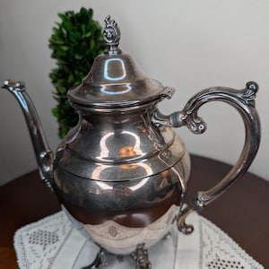 May include: A silver-toned metal teapot featuring a curved spout, a decorative handle, and a lid with a finial top. The teapot stands on three ornate feet and has a polished, reflective surface that shows light reflections and a rounded body shape.