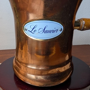 May include: A copper-colored, flared-top saucepan with a wooden handle. The saucepan has a light blue oval label that reads "Le Saucier" in script. The pot sits on a dark wooden base.