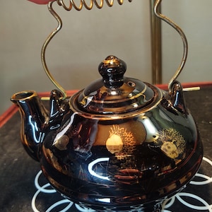 May include: A black ceramic teapot with a gold trim and a spring-loaded handle. The teapot has a floral design and is sitting on a white crocheted coaster.