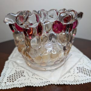 May include: A glass bowl featuring a textured surface of raised, rounded bubbles. The upper section displays vibrant magenta-colored glass bubbles, while the lower portion consists of clear, amber-tinted glass bubbles with a ruffled, uneven rim.
