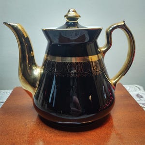 May include: Black teapot with gold trim and a gold lid. The teapot has a decorative design around the middle.