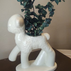 May include: White ceramic lamb planter with a green leafy plant inside. The lamb is standing on all four legs and has a textured surface.