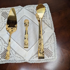 May include: A set of three gold-toned metal serving utensils featuring embossed rose patterns on the handles. The set includes a cake server, a small pair of sugar tongs, and a larger pair of serving tongs, all with a polished, reflective finish.