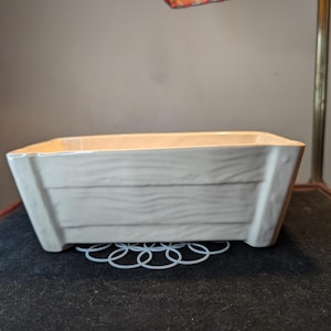 May include: A rectangular, off-white ceramic planter with a wood-grain texture. The planter sits on a white, looped metal stand. The planter is under a lamp with a tan shade and red stitching.