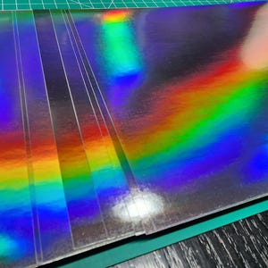 May include: A stack of holographic paper sheets. The sheets have a reflective, iridescent surface that displays a spectrum of colors, including red, orange, yellow, green, blue, and purple. The paper is on a green cutting mat.