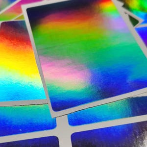 May include: Close-up of a stack of rectangular stickers with a holographic, rainbow-colored finish. The stickers have a white border and reflect a spectrum of colors, including red, yellow, green, and blue.