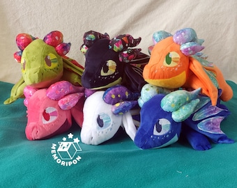 Puede incluir: Seis juguetes de peluche de dragón en varios colores y patrones. Los dragones están dispuestos en semicírculo sobre una superficie verde.