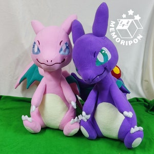 Puede incluir: Dos peluches, una rosa y otra morada, están sentadas sobre una superficie verde. La peluche rosa tiene un ala verde y turquesa y una barriga de color amarillo claro. La peluche morada tiene un ala turquesa y una barriga de color amarillo claro. Ambas peluches tienen ojos grandes y una expresión amigable.