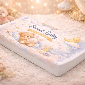 May include: A rectangular baby mattress with a soft, pastel design. The top features a teddy bear and bunny hugging, clouds, a rainbow, and the words "Sweet Baby". The mattress is surrounded by a soft, white surface.
