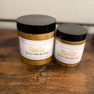 May include: Two jars of turmeric intimate scrub, one larger than the other. Both jars are labeled "Pretty Essentials" and have a brown sugar-like consistency.