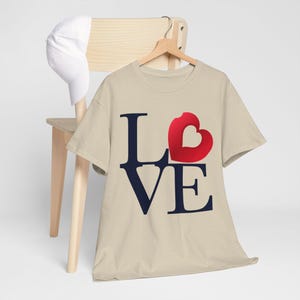 Pode incluir: Uma camiseta bege com um gráfico "LOVE" azul marinho. O "O" de LOVE é substituído por um coração vermelho.