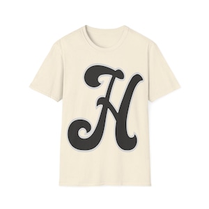 May include: A cream colored t-shirt with a black stylized letter H printed on the front.