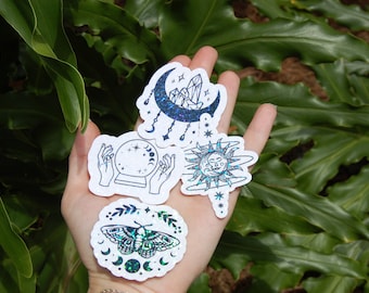 May include: Four iridescent holographic stickers with celestial and mystical themes. The stickers feature a crescent moon with crystals, a crystal ball with hands, a sun with a face, and a moth with a crescent moon.