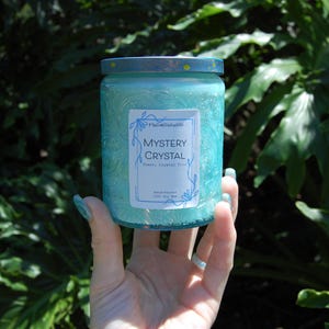 Peut inclure: Une bougie turquoise dans un bocal en verre avec un couvercle bleu. La bougie porte une étiquette indiquant "Mystery Crystal" avec des motifs floraux. La bougie est tenue dans une main devant un fond de feuillage vert.