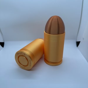 May include: Two 3D-printed bullet-shaped objects. One is upright, with a brown tip and a gold-colored base. The other is lying on its side, also gold-colored, with the text "LUGER 9x19mm" on the base. The objects are on a white background.