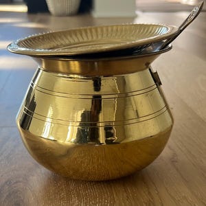 May include: A gold metal pot with a lid and a small ladle. The pot has a round body and a flared rim. The lid has a decorative pattern.
