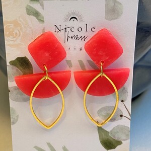 Puede incluir: Un par de pendientes rojos con detalles dorados. Cada pendiente presenta una parte superior cuadrada, una forma de semicírculo y un colgante dorado en forma de lágrima. Los pendientes se exhiben en una tarjeta con el texto "Nicole Thomas Designs".
